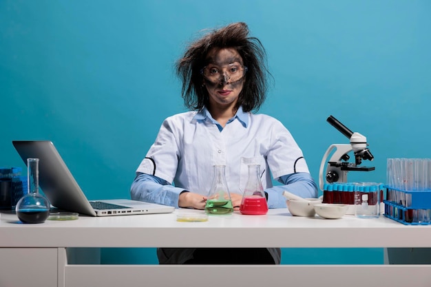 Silly looking crazy chemist with messy hair and dirty face sitting at desk after failed laboratory experiment explosion. Foolish lab worker with wild appearance sitting on blue background. Studio shot