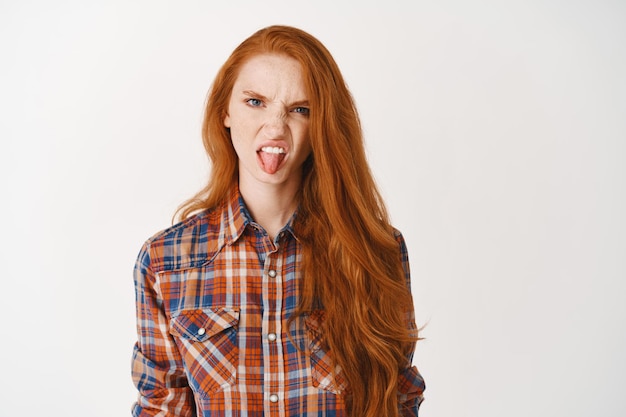 Silly teenage girl with red hair, frowning and showing tongue, pouting disappointed, standing over white wall