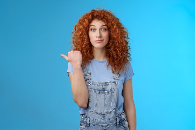 Silly young happy cute redhead woman curly hairstyle smirking rate fine place hang out pointing left thumb introduce not bad choice asking out friend sit nearby cafe standing blue background.