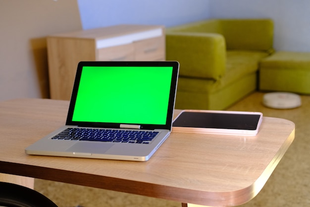 Silver laptop computer with chromakey green display and tablet in the room behind a robot vacuum