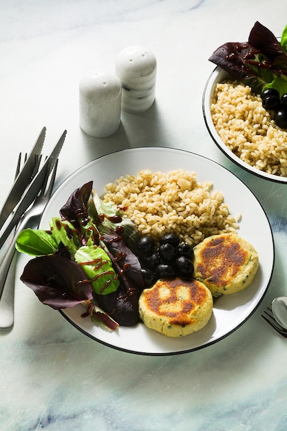 A simple vegan lunch or dinner for a family of two adults and a child brown rice with soy okara and potato patties with fresh salad and olives on a marble table