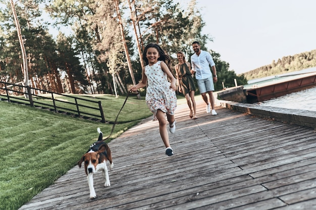 Simply happy. Full length of cute little girl running with dog and smiling while her parents walking behind