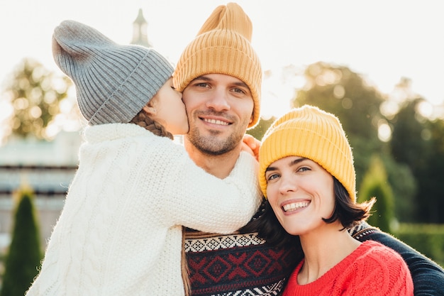 Sincere emotions. Little cute girl in knitted hat and white warm sweater kisses her father with love