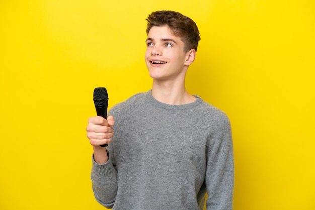 Singer Teenager man picking up a microphone isolated on yellow background looking up while smiling