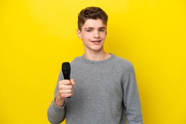 Singer Teenager man picking up a microphone isolated on yellow background smiling a lot