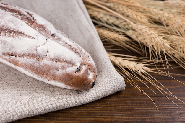 Single loaf of rye bread baguette on brown cloth next to dried wheat stalks over wooden table