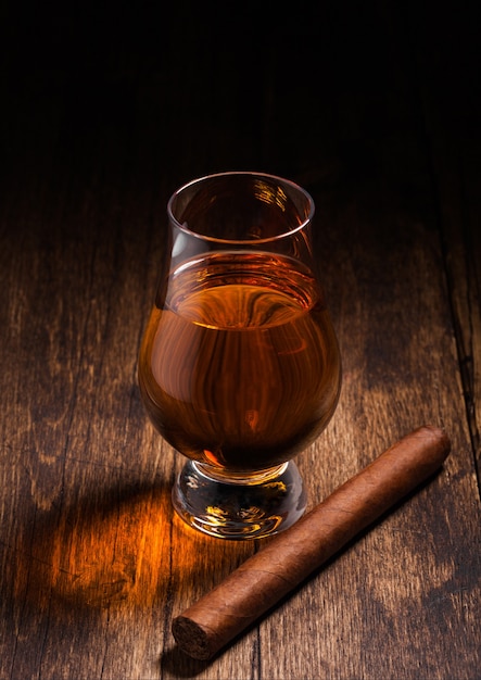 Single malt scotch whiskey in glencairn glass with cuban cigar on wooden table surface. Top view