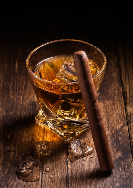 Single malt scotch whiskey in modern crystal glass with ice cubes and cuban cigaron wooden table background. Top view