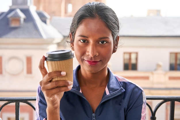 Sipping hot coffee young Indian woman sits on windowsill