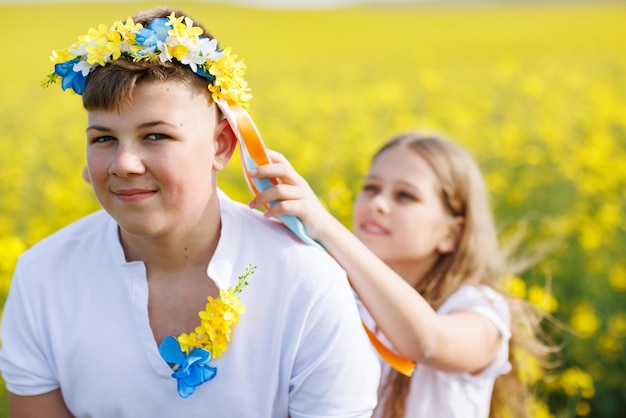 Sister braids ribbons in Ukrainian wreath on head of her teenage brother against backdrop of fields and sky