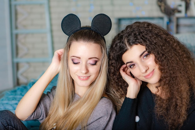 Sisters in warm sweaters with decor at their heads sitting on a plaid and looks at the camera on the background of New Year's decorations