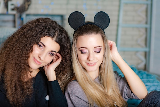 Sisters in warm sweaters with decor at their heads sitting on a plaid and looks at the camera on the background of New Years decorations