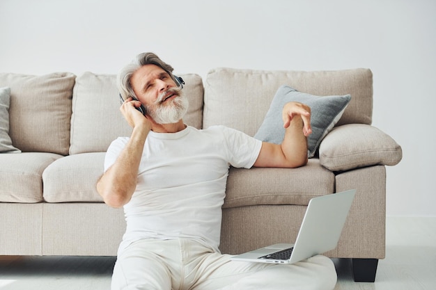 Sits on the ground near sofa with laptop and listens music Senior stylish modern man with grey hair and beard indoors