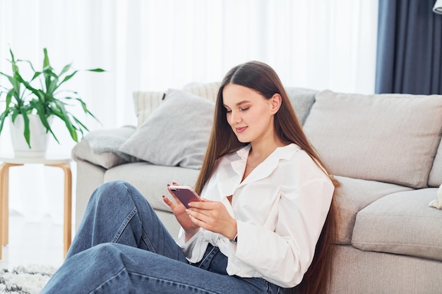 Sits near sofa with phone in hands Young woman in white shirt and jeans is at home