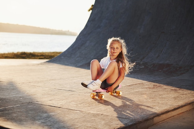 Sits on the skate. Sunny day. Kid have fun at the ramp. Cheerful little girl.