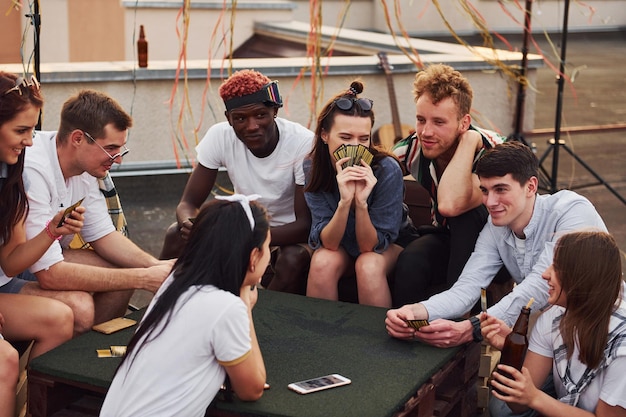 Sitting by table and playing card game Group of young people in casual clothes have a party at rooftop together at daytime