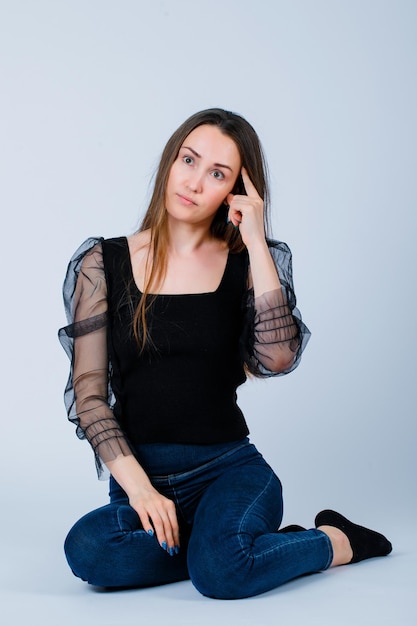 Sitting on flooe girl is thinking by holding forefinger on temple on white background