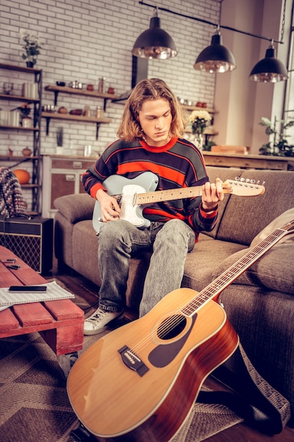 Sitting on sofa. Young blonde-haired student wearing jeans playing the guitar sitting on sofa