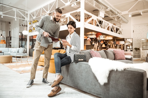 Photo sitting with smartphone. bearded man in grey sweater displaying interested sample while his wife sitting with crossed legs