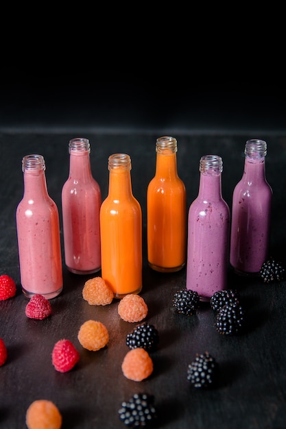 Six bottles with smoothies and raspberry, (red, yellow, blackberry) on black background. Milk shake in glass jar with berries. diet or vegan food concept, fresh vitamin.