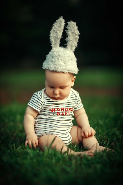 a six-month-old baby sits on the grass in the sunlight in a bunny hat with beautiful ears