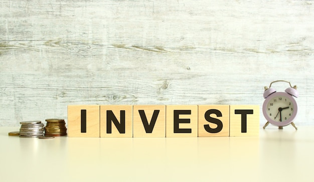 Six wooden cubes with letters are on the table next to the coins. The word is INVEST. On a gray background.