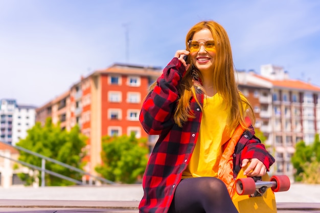 Skater woman in a yellow t-shirt, red plaid shirt and sunglasses, sitting with skateboard in the city, calling with the phone