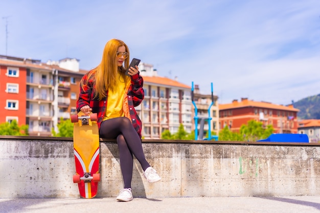 Skater woman in a yellow t-shirt, red plaid shirt and sunglasses, sitting with skateboarding in the city sending an audio message with the phone