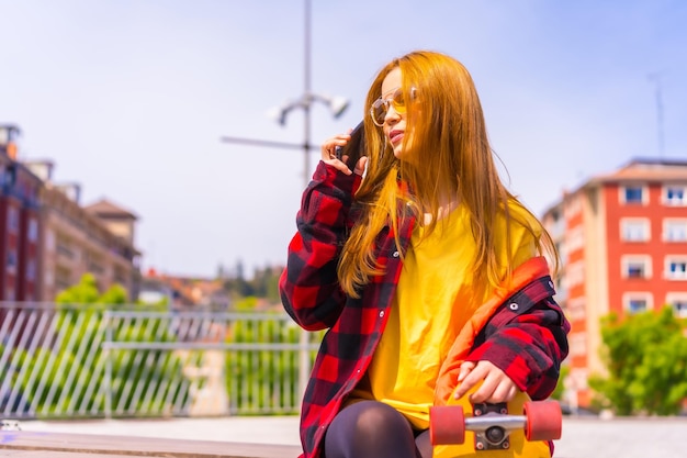 Skater woman in a yellow tshirt red plaid shirt and sunglasses sitting with skateboard in the city calling with the phone