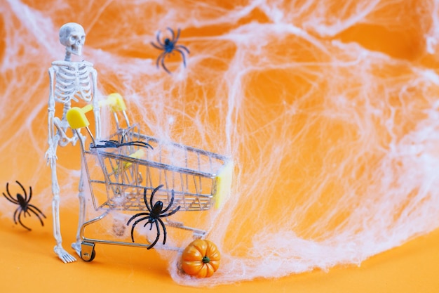 Skeleton with a pumpkin near the trolley spider webs and spiders on an orange background Halloween shopping concept