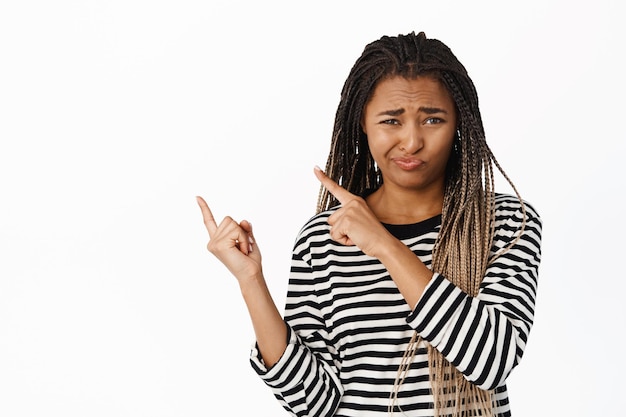 Skeptical african girl expresses doubt points finger away at logo or banner grimaces hesitant or unamused white background