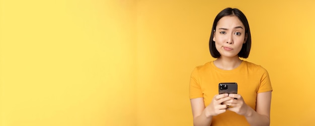 Skeptical asian woman holding smartphone looking with doubt at camera standing over yellow background