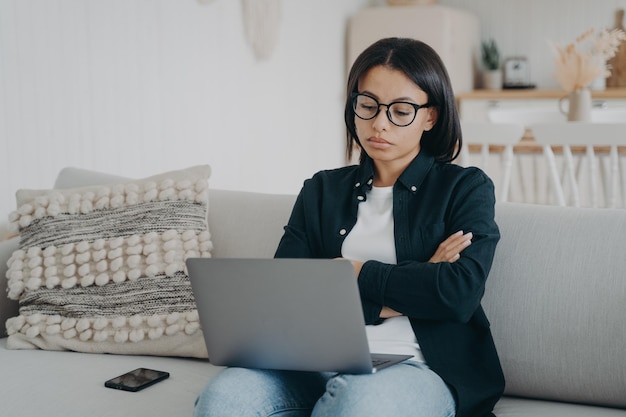 Skeptical businesswoman working on laptop reading email crossed arms sitting on couch at home