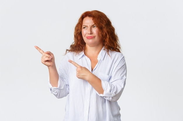 Skeptical and disappointed redhead middle-aged woman, mother dislike promo banner, pointing and looking upper left corner and grimacing judgemental, standing white wall.