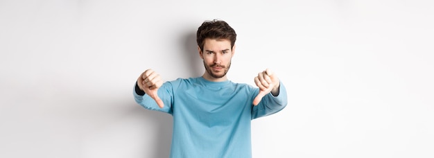 Skeptical handsome guy with beard showing thumbs down in disapproval dislike something bad standing