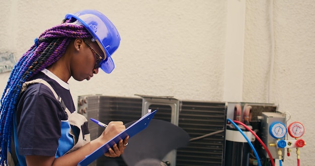 Skilled african american expert commissioned for outdoor air conditioner annual check up, writing findings on clipboard. Seasoned wireman doing hvac system investigation of damaged parts