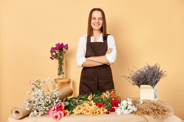 Skilled florists at work Professional floral designers Organic flower market Confident smiling brown haired woman florist standing with crossed arms at her workplace against beige wall