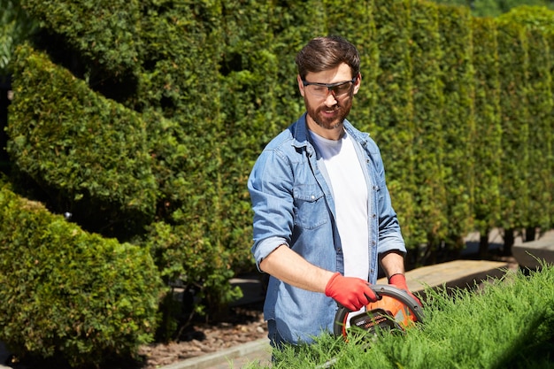 Photo skilled male gardener in protective glasses cutting greenery with hedge trimmer in topiary park