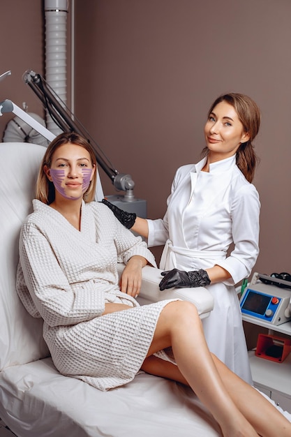 A skilled master cosmetologist stands next to a young woman with tapes on her face and neck to restore facial skin