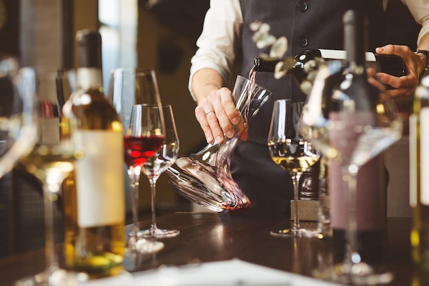 Skilled wine expert pours delicious red beverage into decanter near wineglasses at table