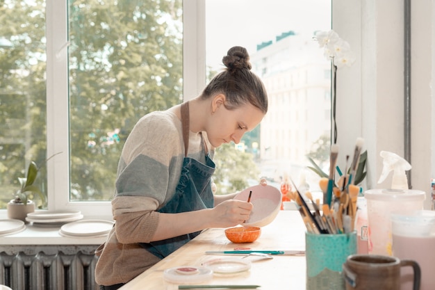 Skilled young woman in apron sitting at table and drawing on ceramic bowl in pottery workshop