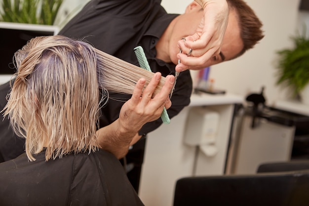 Skillful hairdresser cutting woman blonde hair in beauty salon