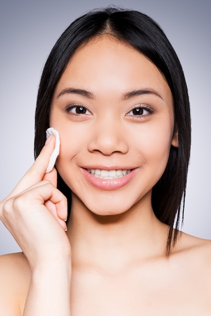 Skin care. Portrait of cheerful young and shirtless Asian woman smiling at camera and touching face with sponge while standing against grey background