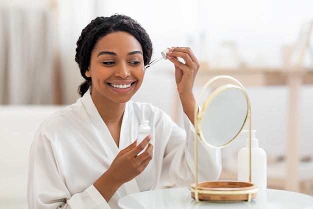 Skincare concept beautiful black lady applying face serum near mirror at home