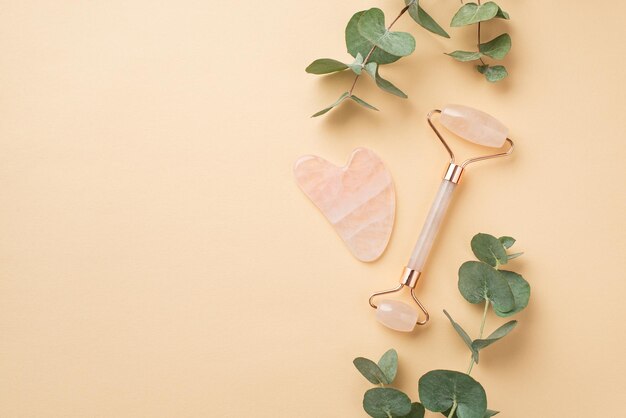 Skincare procedure concept Top view photo of eucalyptus rose quartz roller and gua sha massager on isolated pastel beige background with empty space