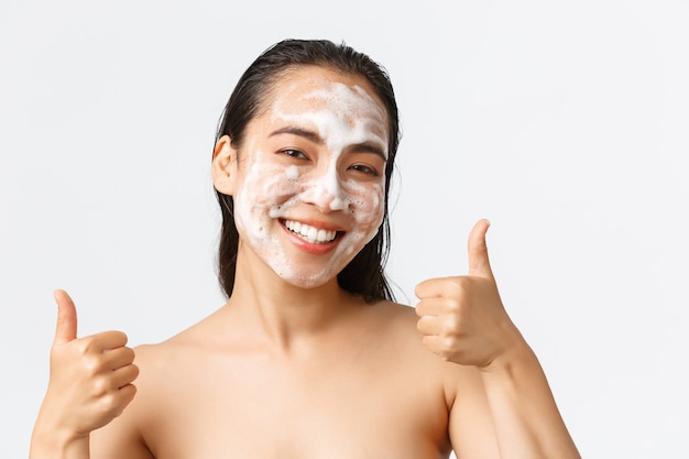 Skincare, women beauty, hygiene and personal care concept. Close-up of satisfied happy, smiling asian woman standing naked and showing thumbs-up while using cleansing foam on face.