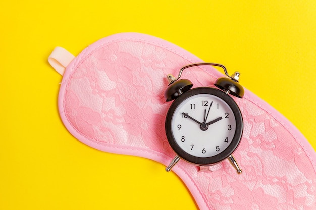 Sleeping eye mask, alarm clock isolated on yellow table