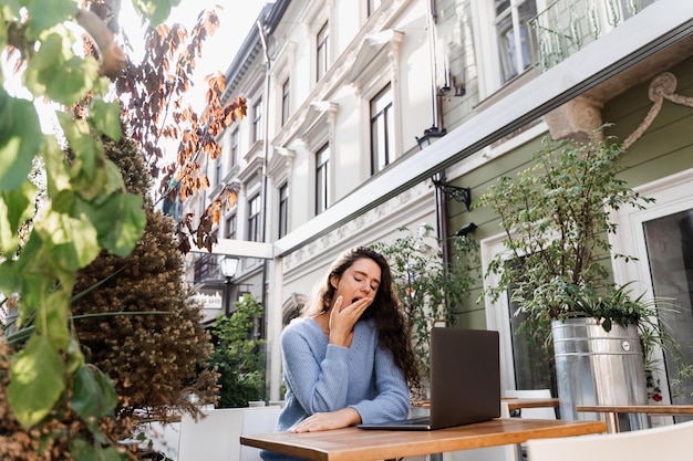 Sleeping girl with laptop is yawning outdoor on terrace in cafe Tired young woman have a break at work and sleeping and relaxing at laptop on workplace due to overtime work