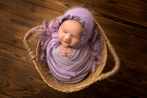 Sleeping newborn baby girl on a background of natural brown wood in a wicker basket A newborn baby with a lilacviolet cap on his head Beautiful portrait of a newborn 12 weeks 14 days old