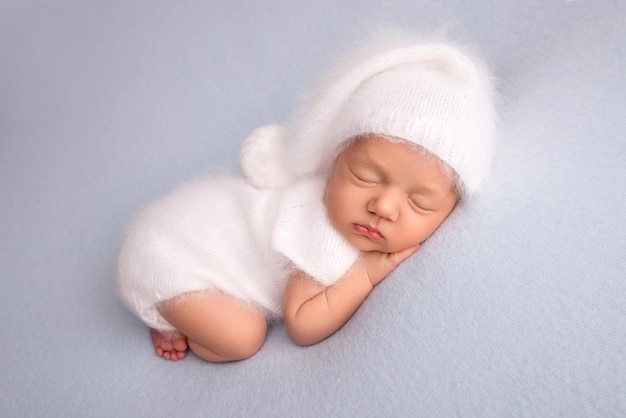 Sleeping newborn girl in the first days of life in a white soft bodysuit with a knitted woolen white cap on a blue background. Studio macro photography, portrait of a newborn. Woman's happiness.